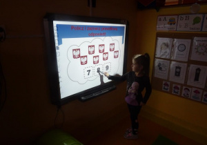 Dziewczynka stoi pod tablicą interaktywną i wskazuje liczbę określającą ilość obrazków znajdujących się na obrazku.